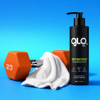 A black pump bottle of GLO Fuel Body Moisturizer stands on a bright blue studio background next to an orange hexagonal dumbbell and a white towel. The bottle label shows the brand GLO FUEL and product name Body Moisturizer.
