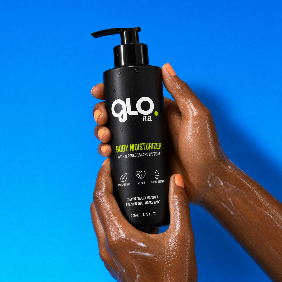 A person's hands holding a black pump bottle of Glo Fuel Body Moisturizer against a vibrant blue background.