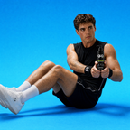 A man in a black athletic tank top, black shorts, and white sneakers sits on a bright blue studio floor, holding a black bottle of GloFyel body moisturizer product. The background is a solid, clean blue.