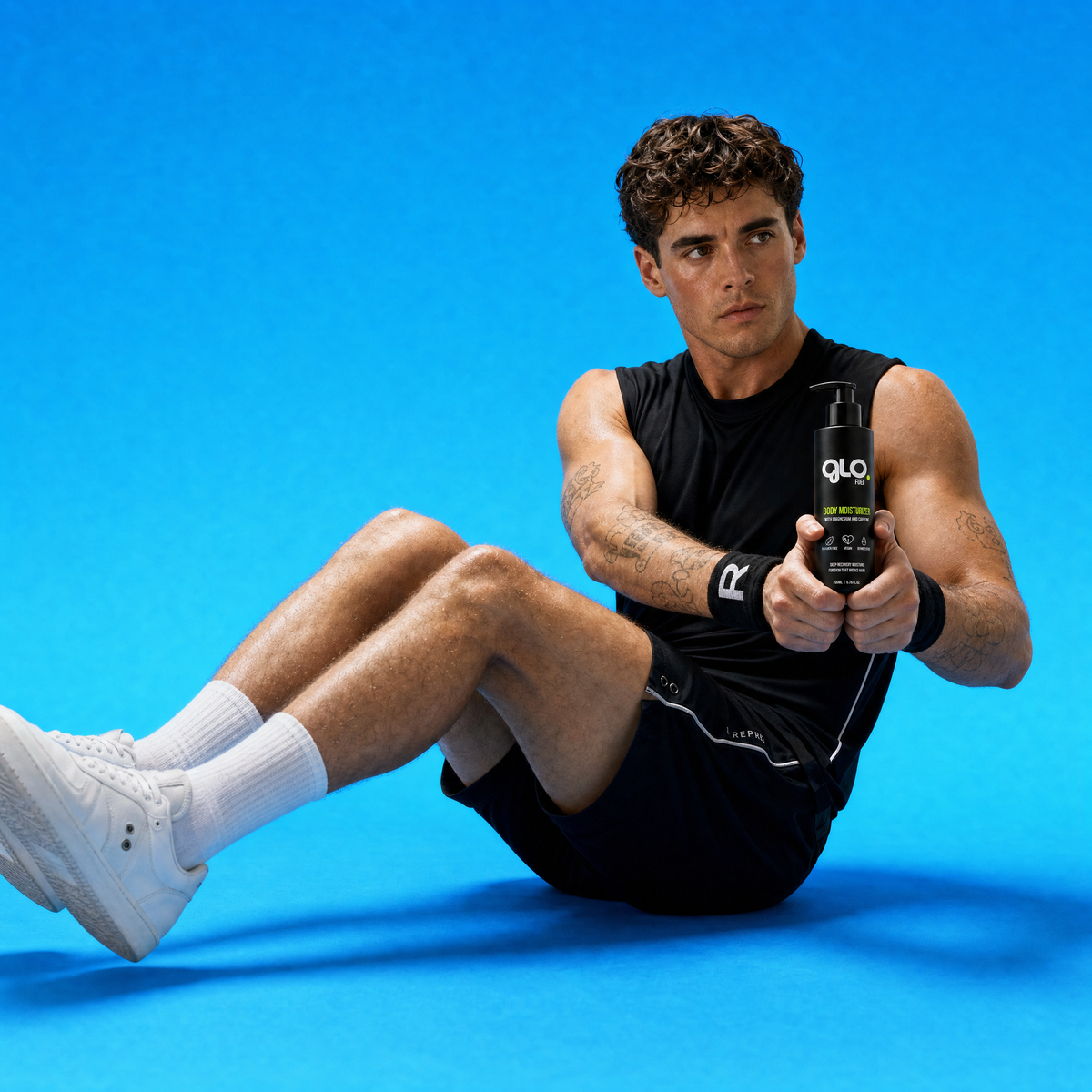 A man in a black athletic tank top, black shorts, and white sneakers sits on a bright blue studio floor, holding a black bottle of GloFyel body moisturizer product. The background is a solid, clean blue.