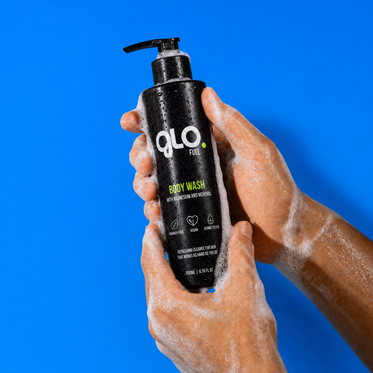 A person's hands covered in white foam hold a black pump bottle of GLO Fuel Body Wash against a solid blue background.