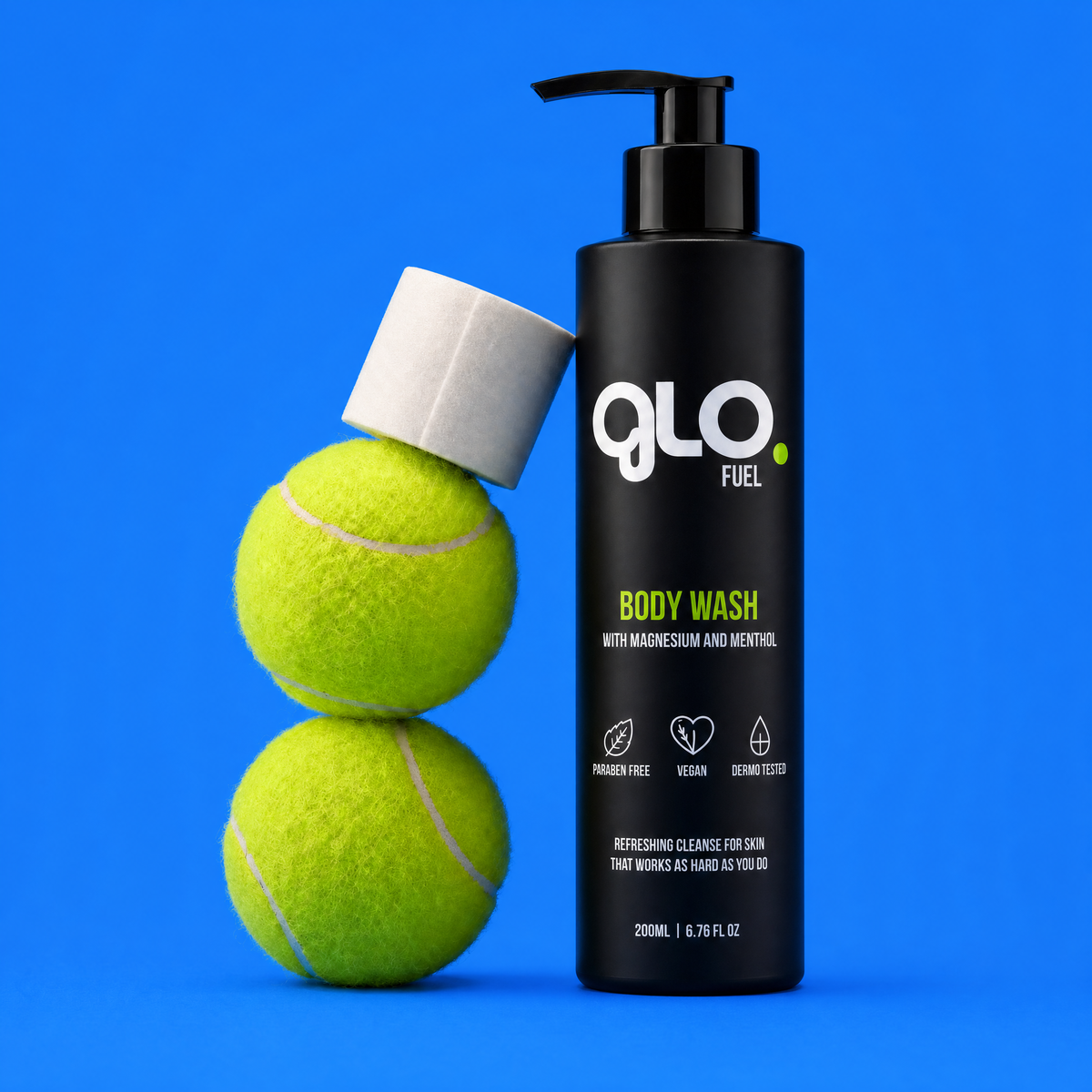 A black pump bottle of Glo Fuel body wash stands on a bright blue background next to two stacked yellow-green tennis balls, with a white cap resting on top of the balls.